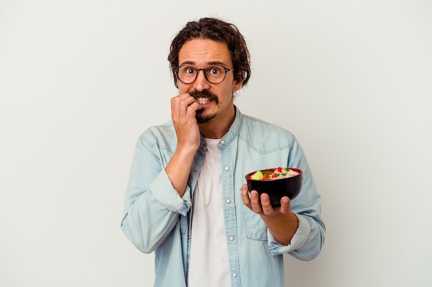 Joven caucásico comiendo un ramen aislado en blanco mordiéndose las uñas, nervioso y muy ansioso.