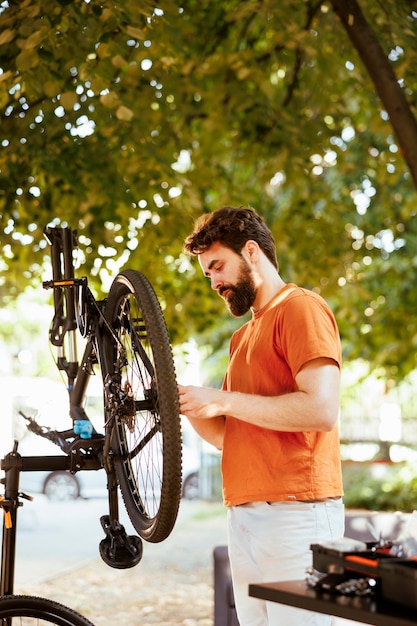 Foto joven caucásico entusiasta afuera haciendo mantenimiento y mantenimiento anual de bicicletas modernas. ciclista masculino sano visto al aire libre revisando la bicicleta en busca de daños para reparar y ajustar.