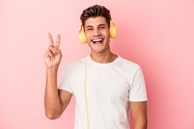 Foto joven caucásico escuchando música aislada sobre fondo rosa alegre y despreocupado mostrando un símbolo de paz con los dedos.