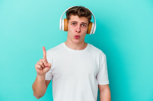 Joven caucásico escuchando música con auriculares aislado sobre fondo azul con una gran idea, concepto de creatividad.