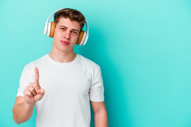 Joven caucásico escuchando música con auriculares aislado sobre fondo azul mostrando el número uno con el dedo.