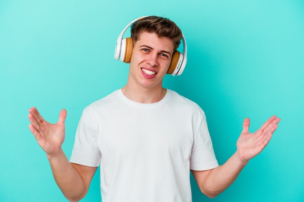 Joven caucásico escuchando música con auriculares aislados en la pared azul recibiendo una agradable sorpresa, emocionado y levantando las manos.