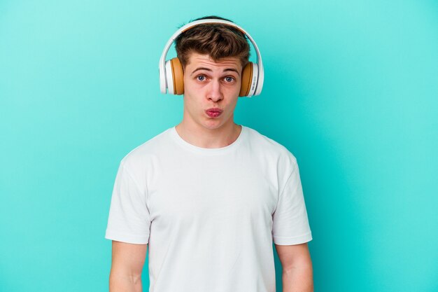 Joven caucásico escuchando música con auriculares en azul se encoge de hombros y abre los ojos confundidos.