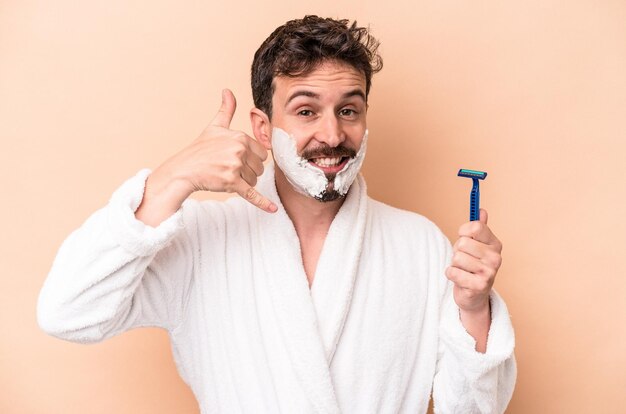 Joven caucásico con espuma de afeitar y sosteniendo una hoja de afeitar aislada en un fondo beige mostrando un gesto de llamada de teléfono móvil con los dedos