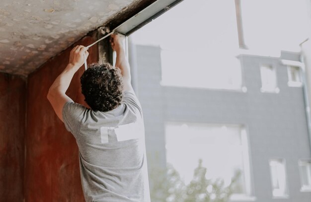 Un joven caucásico está reparando la apertura de una ventana quitando el aislamiento de silicona