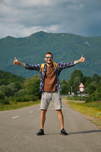 Foto joven caucásico haciendo autostop de pie en la carretera rural vacía en un día soleado de verano