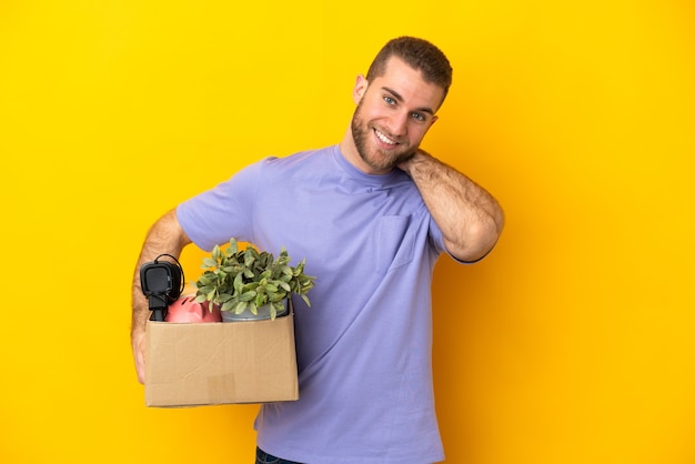 Joven caucásico haciendo un movimiento mientras recoge una caja llena de cosas aisladas en amarillo riendo