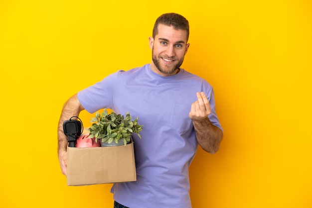 Joven caucásico haciendo un movimiento mientras recoge una caja llena de cosas aisladas en la pared amarilla haciendo gesto de dinero