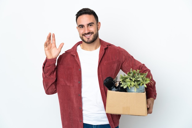 Joven caucásico haciendo un movimiento mientras recoge una caja llena de cosas aisladas sobre fondo blanco saludando con la mano con expresión feliz