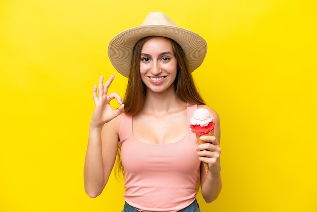 Joven caucásico con un helado de cucurucho aislado de fondo amarillo que muestra el signo de ok con los dedos