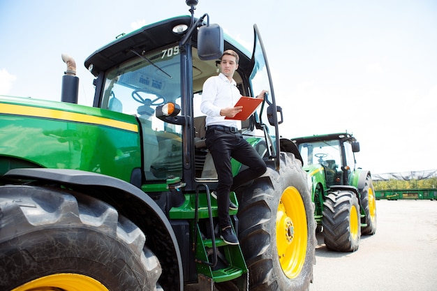 Joven caucásico, inversor, comprador con documentos en tractor, combinar durante el trabajo. Parece seguro, colores de verano brillantes, sol. Agricultura, exposición, maquinaria, venta de producción vegetal