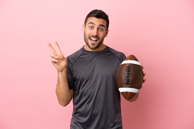 Joven caucásico jugando al rugby aislado sobre fondo rosa sonriendo y mostrando el signo de la victoria