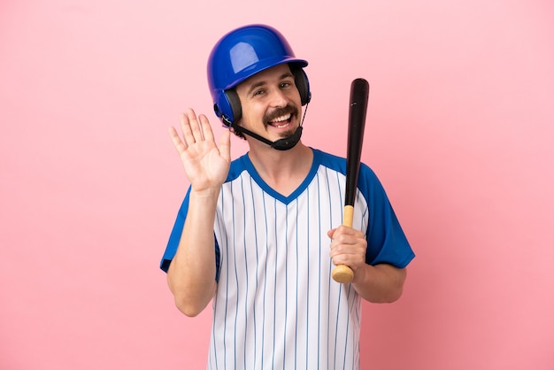 Joven caucásico jugando béisbol aislado sobre fondo rosa saludando con la mano con expresión feliz