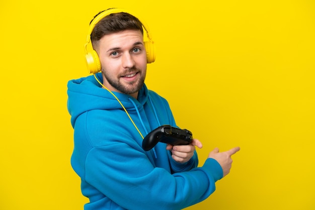 Joven caucásico jugando con un controlador de videojuegos aislado en un fondo amarillo apuntando hacia atrás