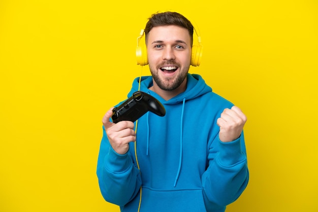 Joven caucásico jugando con un controlador de videojuegos aislado en un fondo amarillo celebrando una victoria en posición de ganador