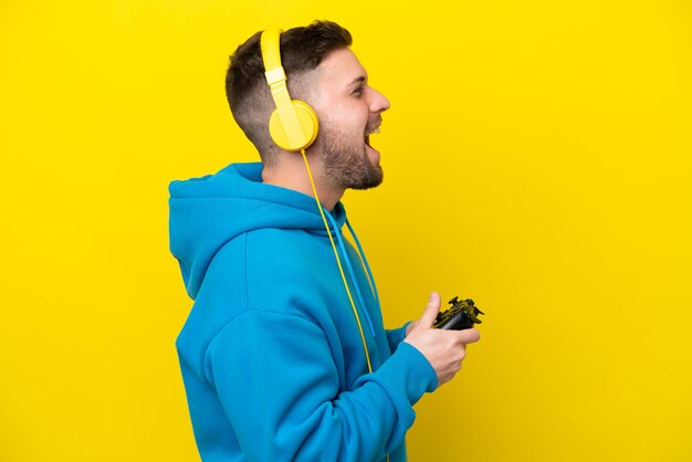 Joven caucásico jugando con un controlador de videojuegos aislado de fondo amarillo riéndose en posición lateral