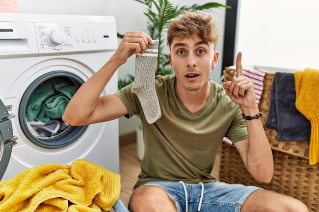 Joven caucásico lavando ropa sosteniendo un calcetín sorprendido con una idea o pregunta señalando con el dedo con la cara feliz número uno