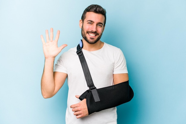 Joven caucásico con la mano rota aislada de fondo azul sonriendo alegre mostrando el número cinco con los dedos