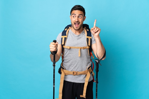 Joven caucásico con mochila y bastones de trekking aislado en azul con la intención de darse cuenta de la solución mientras levanta un dedo