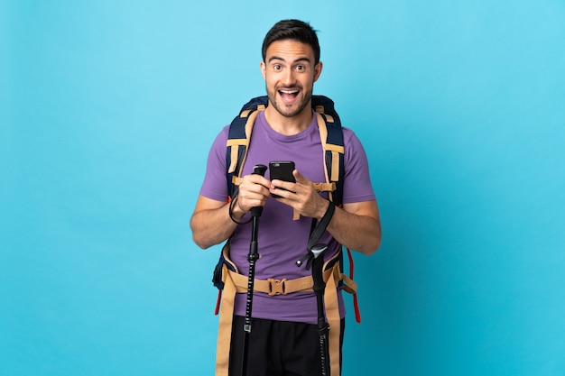 Joven caucásico con mochila y bastones de trekking aislado en la pared azul sorprendido y enviando un mensaje