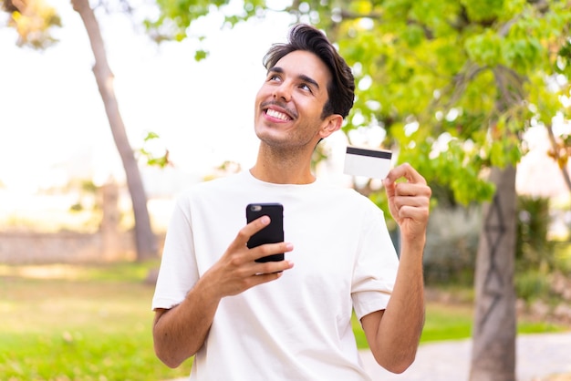 Joven caucásico en un parque comprando con el móvil con tarjeta de crédito mientras piensa