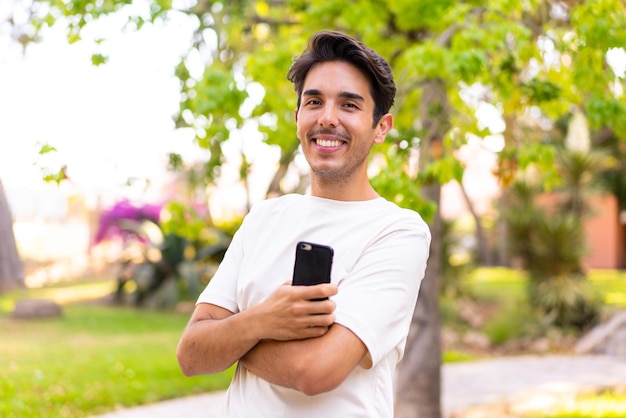 Joven caucásico en un parque sosteniendo un teléfono móvil y con los brazos cruzados