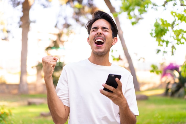 Joven caucásico en un parque usando teléfono móvil y haciendo gesto de victoria