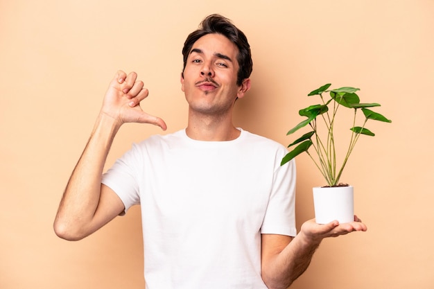 Un joven caucásico que sostiene una planta aislada de fondo beige se siente orgulloso y seguro de sí mismo como un ejemplo a seguir