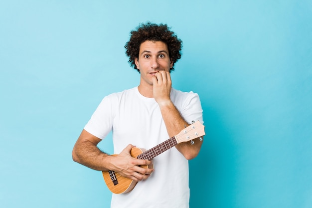 Joven caucásico rizado jugando ukelele morderse las uñas, nervioso y muy ansioso.