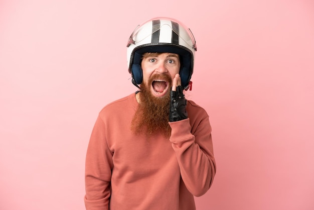 Joven caucásico rojizo con un casco de motocicleta aislado en un fondo rosado con expresión facial sorprendida y sorprendida