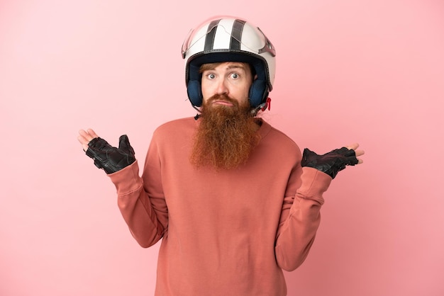 Joven caucásico rojizo con un casco de motocicleta aislado en un fondo rosado que tiene dudas mientras levanta las manos