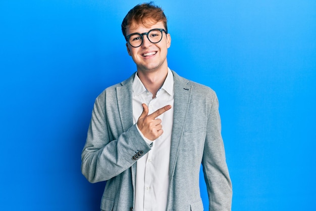 Joven caucásico con ropa elegante y anteojos alegre con una sonrisa en la cara apuntando con la mano y el dedo hacia un lado con expresión feliz y natural en la cara