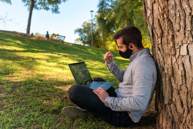 Joven caucásico sentado en el césped mientras mira un partido en la computadora portátil usando su computadora portátil en un hermoso parque en un día soleado.