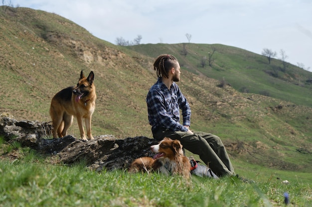 Joven caucásico sentado en una colina con dos perros en primavera y disfrutando de las vistas de la naturaleza