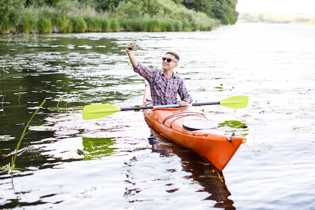 Joven caucásico sentado en un kayak y tomando un selfie en un teléfono móvil El concepto de entretenimiento acuático