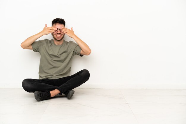 Foto joven caucásico sentado en el suelo aislado sobre fondo blanco cubriéndose los ojos con las manos