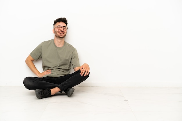 Foto joven caucásico sentado en el suelo aislado sobre fondo blanco posando con los brazos en la cadera y sonriendo