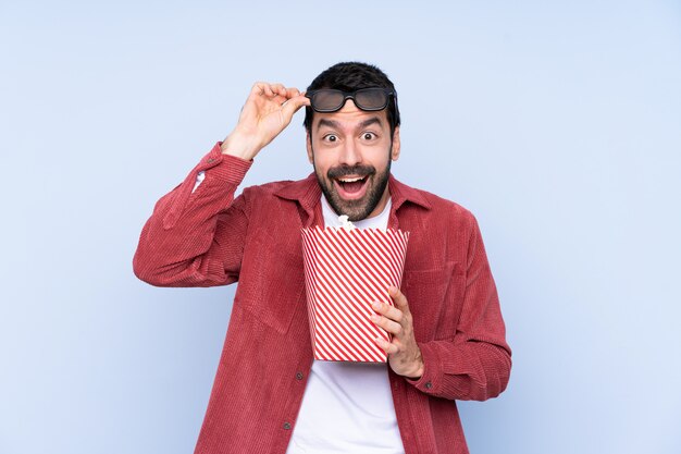 Joven caucásico sobre pared azul sorprendido con gafas 3d y sosteniendo un gran cubo de palomitas de maíz