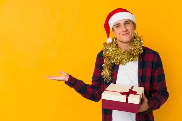 Joven caucásico con sombrero de navidad sosteniendo un presente aislado en la pared amarilla que muestra un espacio de copia en una palma