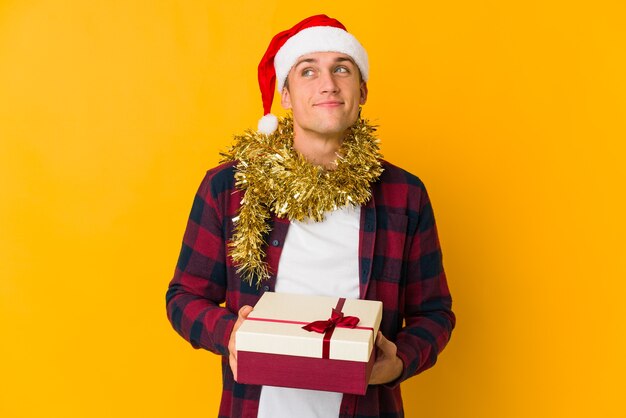 Joven caucásico con sombrero de navidad sosteniendo un regalo aislado en amarillo, soñando con lograr metas y propósitos