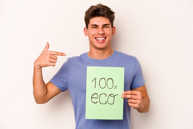 Joven caucásico sosteniendo 100 carteles ecológicos aislados en una persona de fondo blanco señalando con la mano un espacio de copia de camisa orgulloso y confiado