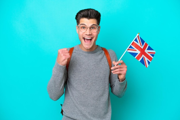 Joven caucásico sosteniendo una bandera del Reino Unido aislada de fondo azul celebrando una victoria en posición de ganador