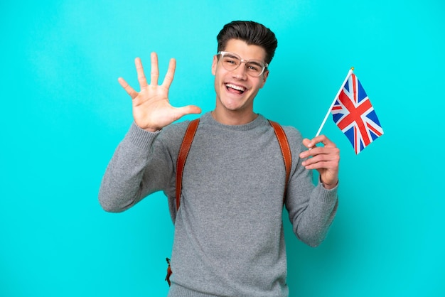Joven caucásico sosteniendo una bandera del Reino Unido aislada de fondo azul contando cinco con los dedos