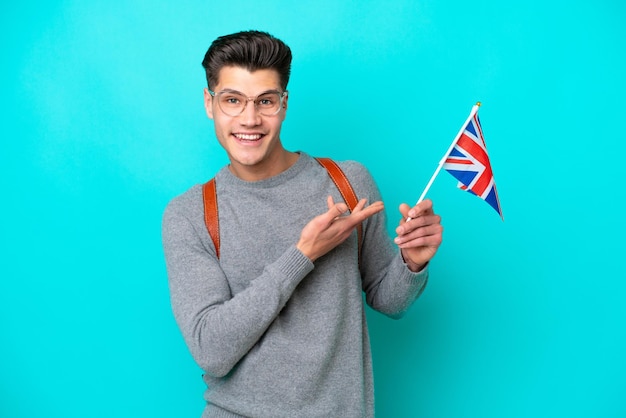 Joven caucásico sosteniendo una bandera del Reino Unido aislada de fondo azul extendiendo las manos a un lado para invitar a venir