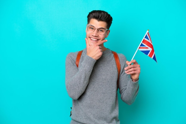 Joven caucásico sosteniendo una bandera del Reino Unido aislada de fondo azul feliz y sonriente