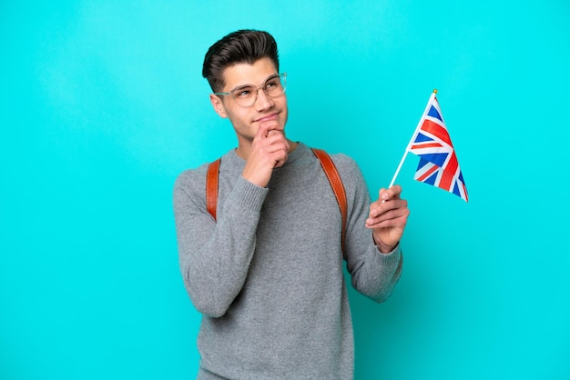 Joven caucásico sosteniendo una bandera del Reino Unido aislada de fondo azul y mirando hacia arriba