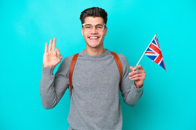 Joven caucásico sosteniendo una bandera del Reino Unido aislada de fondo azul mostrando el signo de ok con los dedos