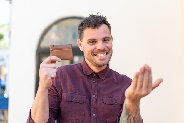 Foto joven caucásico sosteniendo una billetera al aire libre invitando a venir con la mano feliz de que hayas venido