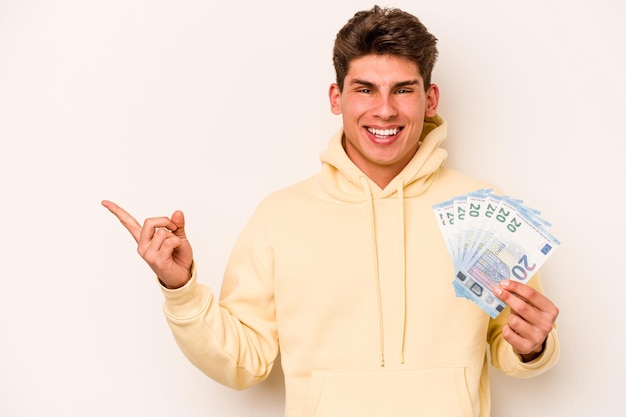 Joven caucásico sosteniendo billetes aislados de fondo blanco sonriendo y señalando a un lado mostrando algo en el espacio en blanco