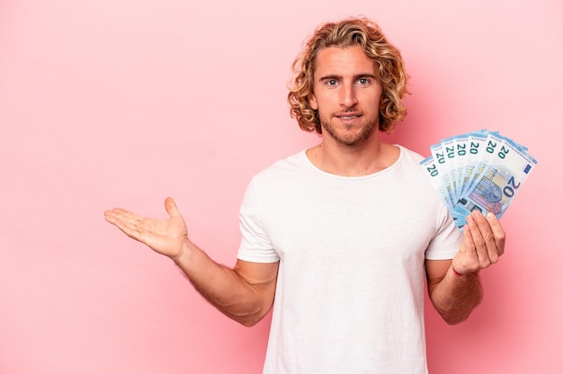 Joven caucásico sosteniendo billetes aislados en un fondo rosado mostrando un espacio de copia en una palma y sosteniendo otra mano en la cintura.